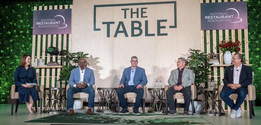 The Table 2025 speakers seated on stage in front of large signage reading "the table" and green leaves on either side. pictured: