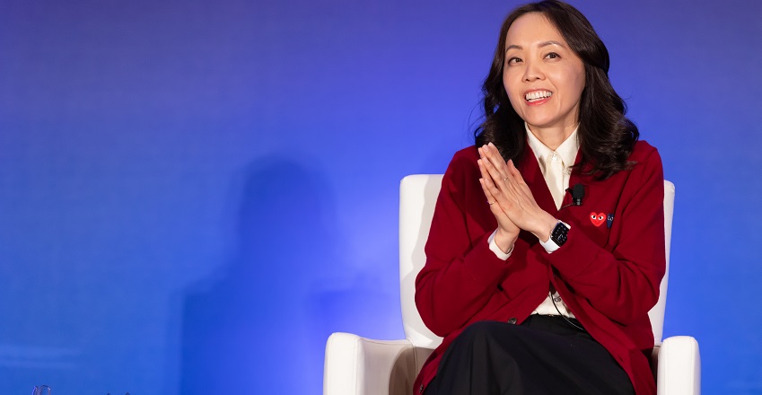 Andrea Cheng at a talk sitting in front of blue screen smiling with hands clasped together wearing red blazer