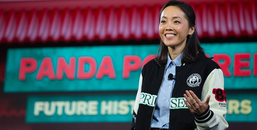 Andrea Cheng in a black vest standing in front of a backdrop picturing a green and red Panda restaurant sign